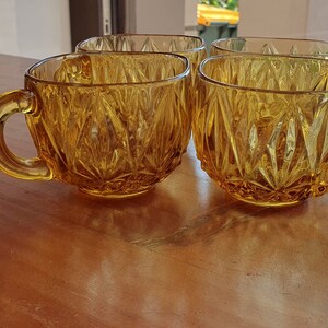 May include: Four amber glass cups with a diamond pattern. The cups have handles and are sitting on a wooden surface.