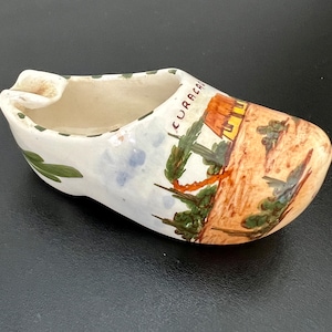 Könnte beinhalten: Ein weißer, keramik-schuhförmiger Aschenbecher mit einem grün, braun und blau bemalten Design. Der Schuh hat das Wort "CURACAO" auf der Seite bemalt.