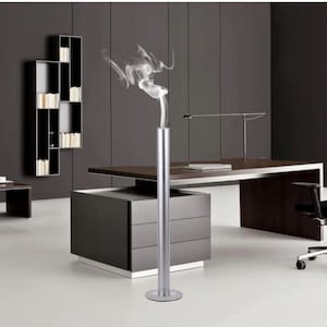 May include: A silver metal diffuser with a cylindrical shape and a round base. The diffuser is emitting a plume of white smoke in a modern office setting.