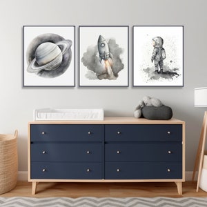 Könnte beinhalten: Drei Schwarz-Weiß-Aquarelle mit Weltraumthema. Die Drucke zeigen einen Planeten mit Ringen, eine Rakete und einen Astronauten. Die Drucke sind in schwarzen Rahmen gerahmt und hängen an einer weißen Wand über einer dunkelblauen Kommode.