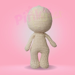 May include: A crocheted doll with a cream-colored body and a round head. The doll is standing on a pink background.