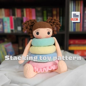 May include: A crocheted stacking toy pattern for a doll. The doll is sitting down with a pink skirt and a stack of four colorful rings around its waist. The rings are light blue, yellow, light blue, and pink. The doll has brown hair and a sweet face.