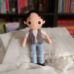 May include: A crocheted doll with black hair, a white shirt, a gray vest, and blue jeans. The doll is standing on a white surface with white pom-poms.