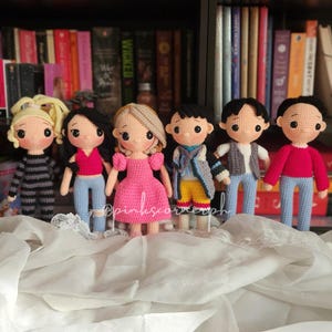 May include: Six crocheted dolls, each with a different outfit and hairstyle. The dolls are standing on a white fabric surface. The dolls are all wearing different colored clothing, including pink, blue, red, and gray.