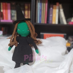 May include: A handmade crochet witch doll with a green face, brown hair, and a black dress and hat. The doll has green hands and feet. The background includes a bookshelf with books and white tissue paper.