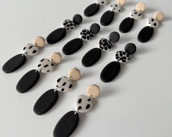 May include: A collection of handmade earrings with a modern design. Each earring features a black oval, a cream-colored circle, and a black and white spotted shape. The earrings are arranged diagonally on a white surface.