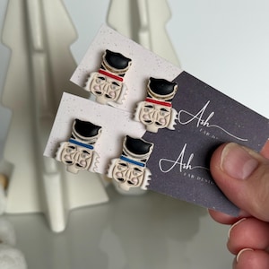 Puede incluir: Cuatro pequeños pendientes de botón con forma de soldaditos de juguete, cada uno con un sombrero negro, cuello rojo o azul y una cara detallada. Los pendientes se muestran en tarjetas blancas, con una tarjeta gris oscuro con el texto "Ash FAB DESIGNS".