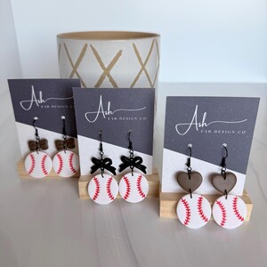 Peut inclure: Boucles d'oreilles sur le thème du baseball avec des balles blanches, des coutures rouges et différents motifs supérieurs. Elles sont présentées sur de petits présentoirs en bois avec des cartes grises et blanches. Texte : "Ash FAB DESIGN CO."