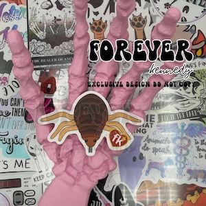 May include: A pink plastic hand with four fingers, holding a brown and black tick sticker. The hand is surrounded by other stickers, including a sticker with the text "FOREVER kennedy EXCLUSIVE DESIGN DO NOT COPY".