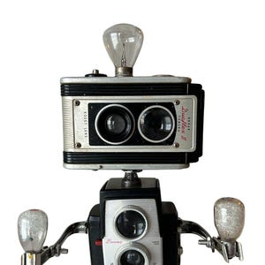 May include: A whimsical robot sculpture crafted from vintage cameras. The head features a Kodak Duaflex II camera with a lightbulb, while the body is a Brownie Starflex camera. Additional lightbulbs serve as arms, creating a unique art piece.