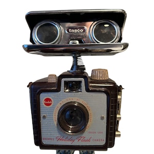 May include: A whimsical sculpture of a vintage Kodak Brownie Holiday Flash camera with Tasco binoculars as eyes. The camera body is brown and silver, with the words "Kodak" and "Holiday Flash Camera" visible. The binoculars are black.