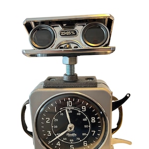 May include: A quirky robot sculpture crafted from vintage components. It features a silver clock face with black numerals and hands, a pair of binoculars, and a metal base. The clock face reads "Franklin" and "Made in Great Britain". The clock face is approximately 10 cm in diameter.
