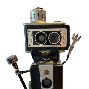 May include: A whimsical assemblage of vintage cameras, creatively arranged to resemble a robot. The robot features a black bow tie, a microphone, and a metallic hand. The cameras are black and silver, with the text "Brownie Hawkeye Camera Flash Model" visible.