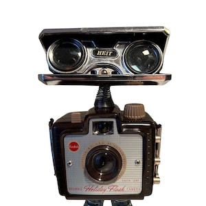 May include: A whimsical robot sculpture crafted from vintage camera parts. The head is made of binoculars, the body is a Kodak Brownie Holiday Flash camera, and the feet are metal. The robot has a spring neck and a brown and silver color scheme.