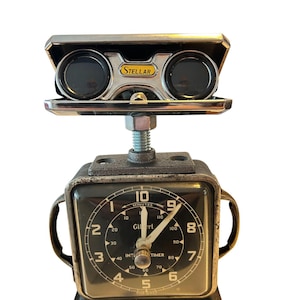 May include: A quirky sculpture combining a vintage clock and binoculars. The black clock face has white numerals and hands, with the word "Gilbert". The binoculars are silver and branded "STELLAR".