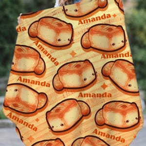 May include: A personalized blanket featuring a repeating pattern of cartoon bread with smiling faces and the name "Amanda" in a playful font. The blanket has a warm, yellow-orange background with star accents.