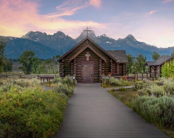 May include: A wooden chapel with a cross on the roof sits in front of a mountain range. The chapel has a wooden door and windows. The mountains are in the background and the sky is a light pink and blue.