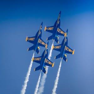 Pode incluir: Quatro jatos de combate azuis e amarelos voam em formação contra um céu azul brilhante, deixando rastros de fumaça branca atrás deles. Cada jato tem "U.S. NAVY" escrito no lado.