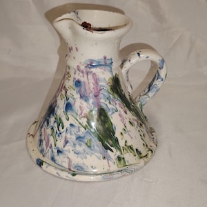 Puede incluir: Una jarra de cerámica blanca con asa y base. La jarra tiene un diseño abstracto colorido con salpicaduras de pintura azul, verde y rosa.