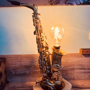 Pode incluir: Uma lâmpada feita com um saxofone vintage. O instrumento de latão apresenta uma grande lâmpada de estilo Edison, emitindo um brilho quente. A lâmpada é montada em uma base de madeira, criando uma luminária única e artística.