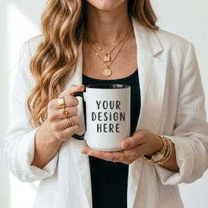 Puede incluir: Taza de cerámica blanca con interior y asa negros, con el texto "YOUR DESIGN HERE" en negro. La taza es sostenida por una persona que lleva una blazer blanca y joyas doradas.