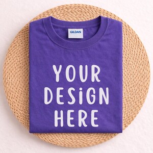 Puede incluir: Una camiseta morada doblada con el texto "YOUR DESIGN HERE" en blanco, colocada sobre una estera redonda tejida. La camiseta tiene cuello redondo y una pequeña etiqueta "GILDAN".