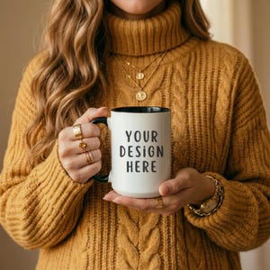 Puede incluir: Taza de cerámica blanca con interior y asa negros, con el texto "YOUR DESIGN HERE" en negro. La taza es sostenida por una persona que lleva un jersey de punto trenzado amarillo mostaza y joyas doradas.