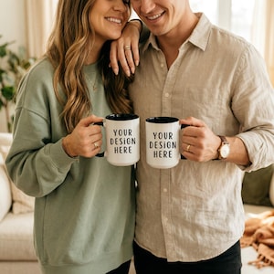 Puede incluir: Dos tazas de café blancas con interior y asas negras. Cada taza tiene el texto "YOUR DESIGN HERE" impreso en negro. Las tazas son sostenidas por dos personas, una con una sudadera verde y otra con una camisa beige.