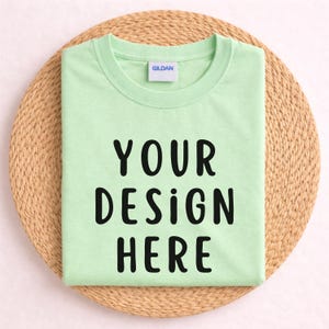 May include: A folded, light green t-shirt with the text "YOUR DESIGN HERE" in black. The shirt is on a round, woven, light brown mat. The Gildan brand label is visible on the collar.