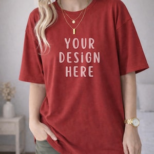 Puede incluir: Una camiseta roja con el texto "YOUR DESIGN HERE" en letras blancas. La camiseta tiene mangas cortas y un corte holgado. Se ve un reloj dorado y collares superpuestos.