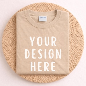 Puede incluir: Una camiseta beige doblada con el texto "YOUR DESIGN HERE" en letras blancas. La camiseta está colocada sobre un salvamanteles redondo y beige tejido. La etiqueta de la marca Gildan es visible en el cuello.