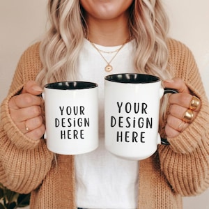 May include: Two white ceramic mugs with black interiors and handles. The mugs have the text "YOUR DESIGN HERE" printed in black. The mugs are held up by a person wearing a tan sweater.