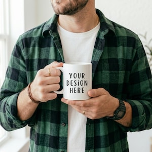 Puede incluir: Una taza de cerámica blanca con el texto "YOUR DESIGN HERE" en negro, sostenida por una persona. La persona lleva una camisa a cuadros verde y negra sobre una camiseta blanca. Se ve una pulsera de cuero marrón y un reloj negro.