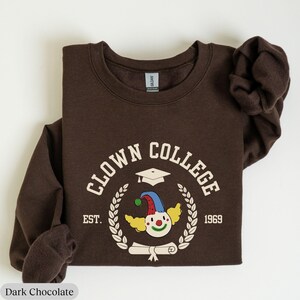 May include: A brown sweatshirt with a white graphic of a clown wearing a graduation cap and holding a diploma. The graphic says "Clown College Est. 1969".
