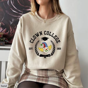 May include: A beige sweatshirt with the text "Clown College" in black lettering, a graduation cap, and a clown face with a red nose. The text "Est. 1969" is also included.