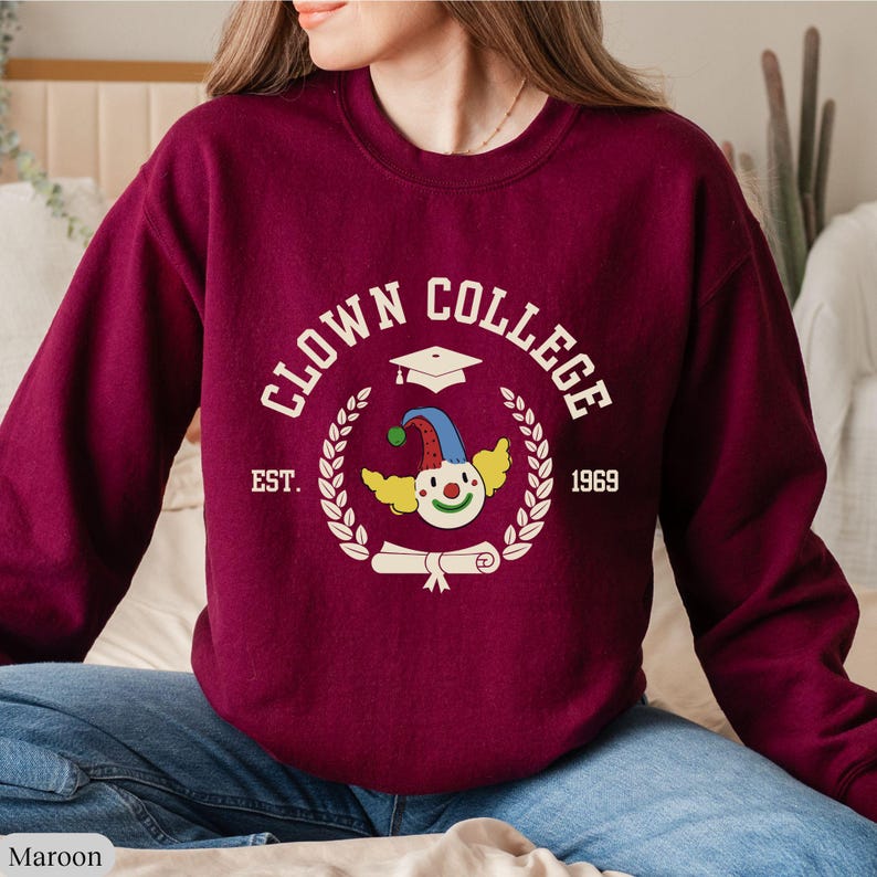 May include: A maroon sweatshirt with a white graphic of a clown wearing a graduation cap and holding a diploma. The graphic also includes the text "Clown College", "Est. 1969", and a wreath of leaves.
