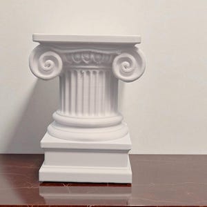 Può includere: Una colonna ionica bianca in miniatura con un capitello dettagliato con volute. La colonna ha un fusto scanalato e poggia su una base quadrata. La colonna è esposta su una superficie marrone-rossastra.