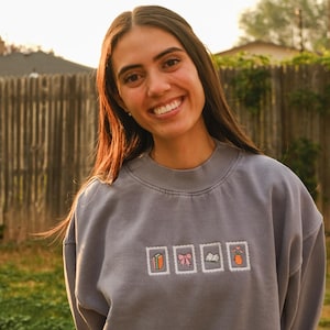 May include: A gray sweatshirt with embroidered postage stamp designs featuring a book, bow, and other objects. The sweatshirt is worn by a person smiling in an outdoor setting.