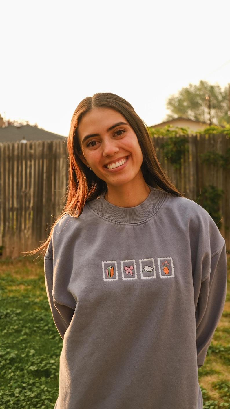 May include: A gray sweatshirt with embroidered postage stamp designs featuring a book, bow, and other objects. The sweatshirt is worn by a person smiling in an outdoor setting.