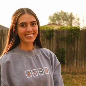 May include: A woman wearing a light blue sweatshirt with embroidered images of a carrot, a gift, a bow, a book, and a carrot. The background includes a wooden fence and greenery.