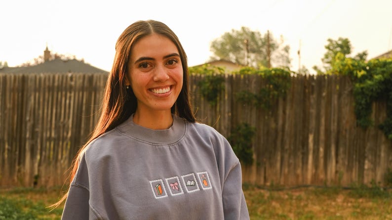 May include: A woman wearing a light blue sweatshirt with embroidered images of a carrot, a gift, a bow, a book, and a carrot. The background includes a wooden fence and greenery.