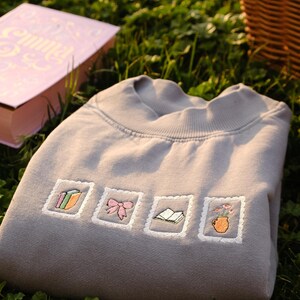 May include: A gray sweatshirt embroidered with four postage stamp-like designs. The designs include a stack of books, a pink bow, an open book, and a vase of flowers. The sweatshirt is folded on green grass, with a book and a picnic basket in the background.