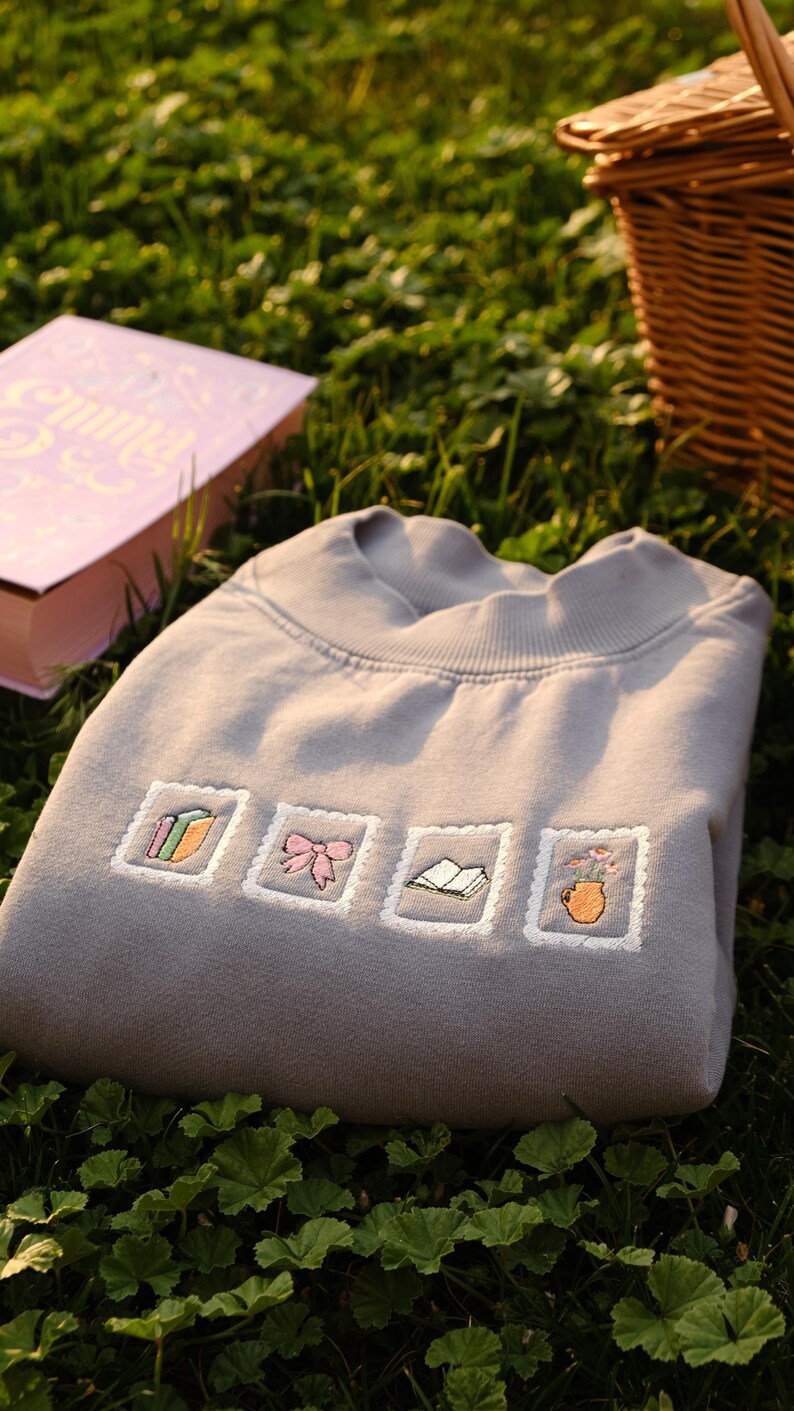 May include: A gray sweatshirt embroidered with four postage stamp-like designs. The designs include a stack of books, a pink bow, an open book, and a vase of flowers. The sweatshirt is folded on green grass, with a book and a picnic basket in the background.