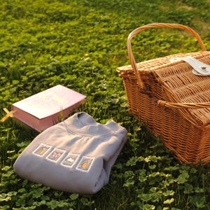 May include: A picnic scene with a woven wicker basket, a gray sweatshirt with embroidered designs, and a pink book with a yellow bookmark, all set on a bed of green grass. The basket has a handle and a white clasp.