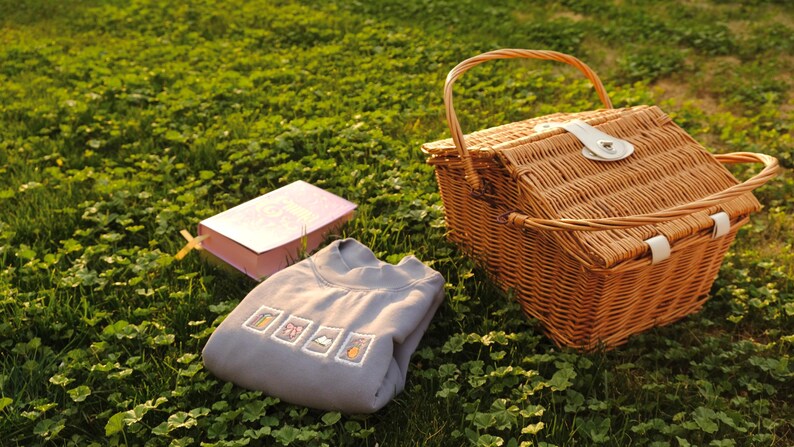 May include: A picnic scene with a woven wicker basket, a gray sweatshirt with embroidered designs, and a pink book with a yellow bookmark, all set on a bed of green grass. The basket has a handle and a white clasp.