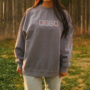 May include: A gray sweatshirt with a crew neck and long sleeves. The sweatshirt features four embroidered postage stamp designs: a pumpkin, a bow, a book, and an acorn. The person is wearing light blue jeans.