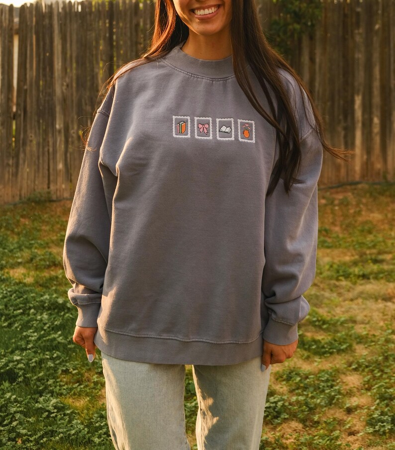 May include: A gray sweatshirt with a crew neck and long sleeves. The sweatshirt features four embroidered postage stamp designs: a pumpkin, a bow, a book, and an acorn. The person is wearing light blue jeans.
