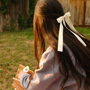May include: A person with long, dark brown hair styled with a white bow. The person is wearing a light purple sweatshirt and holding a woven basket. The background includes a wooden fence and green grass.