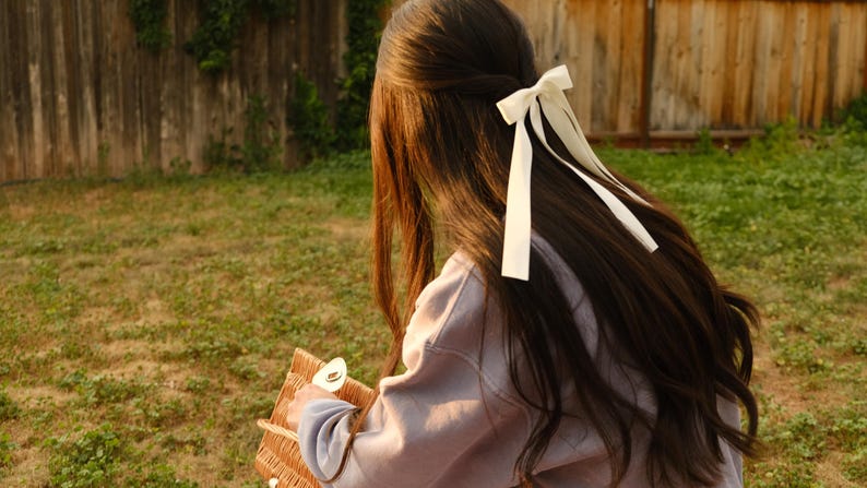 May include: A person with long, dark brown hair styled with a white bow. The person is wearing a light purple sweatshirt and holding a woven basket. The background includes a wooden fence and green grass.