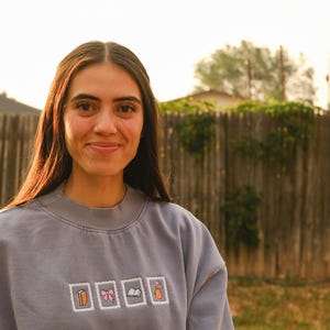 May include: A person wearing a gray sweatshirt with four small, square embroidered designs. The designs include a carrot, a bow, a book, and a bottle. The background features a wooden fence and green foliage.